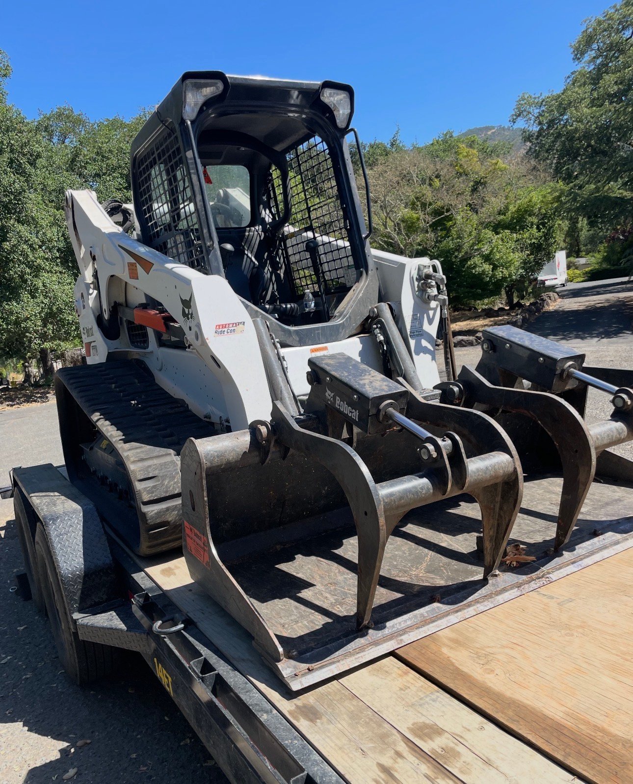 White Bobcat skid steer loader with attachment on flatbed trailer in outdoor setting with trees