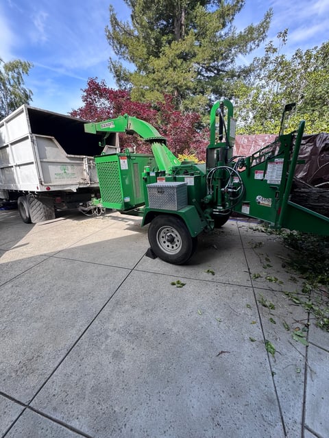 Green wood chipper machine parked next to a truck with trees in background on a driveway